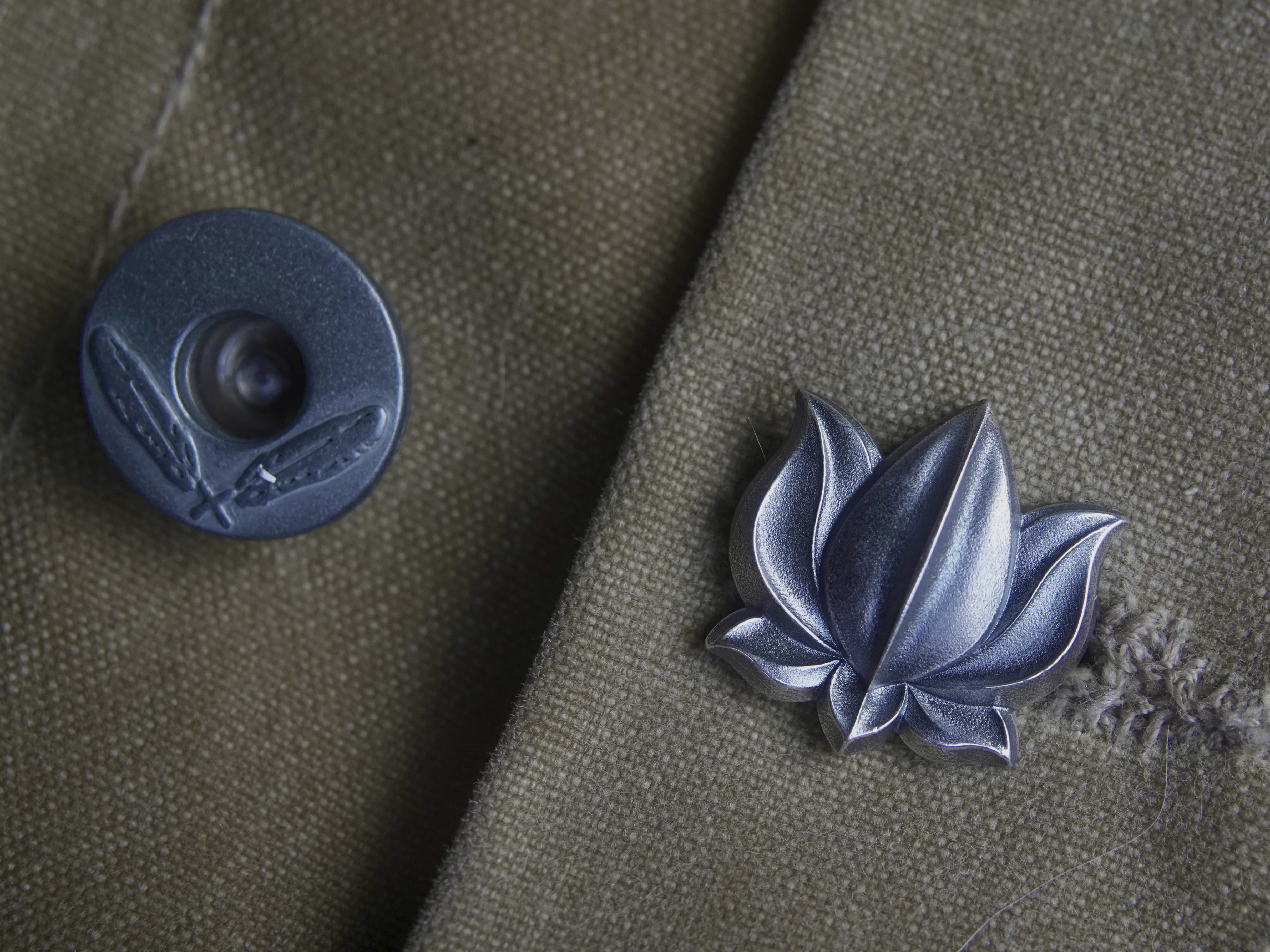 Sterling silver lotus pin on worn olive field canvas beside a metal button